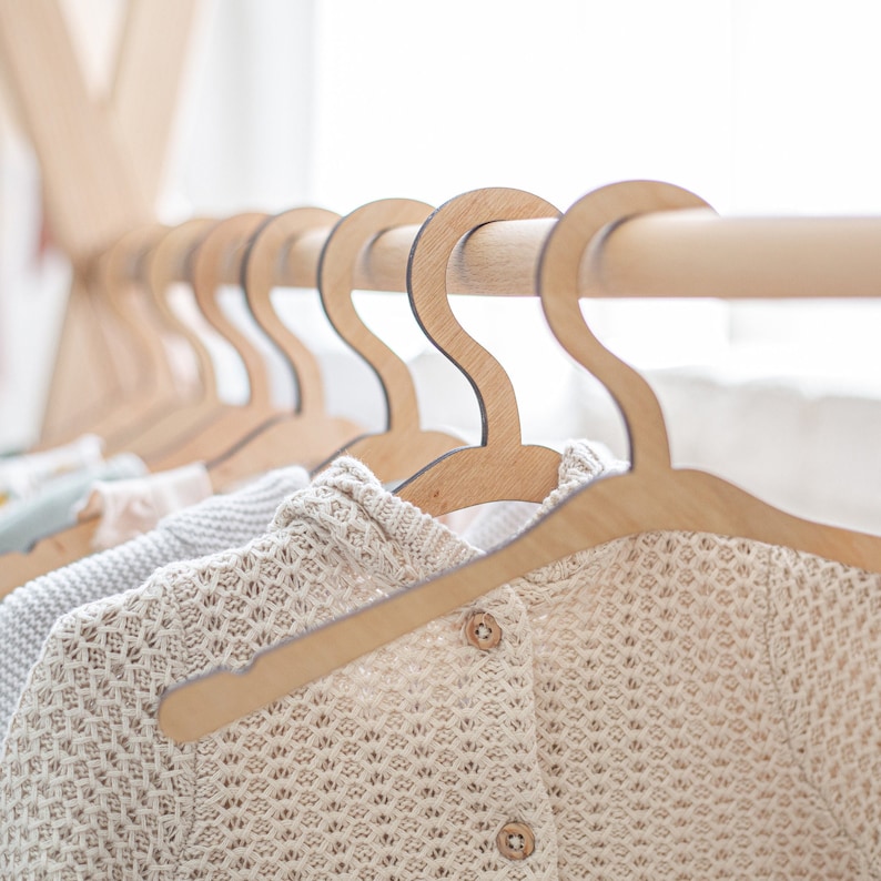 Op de afbeelding: Een close-up van een houten kledingrek met meerdere houten hangers met kleine gebreide kledingstukken. De hangers zijn van natuurlijk hout met een gladde afwerking.