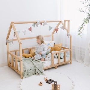May include: A natural wood-framed toddler bed with a house-shaped frame. The bed is decorated with a garland of pastel-colored flags, pillows, and a green blanket. A young child sits inside the bed, surrounded by toys on a white rug.