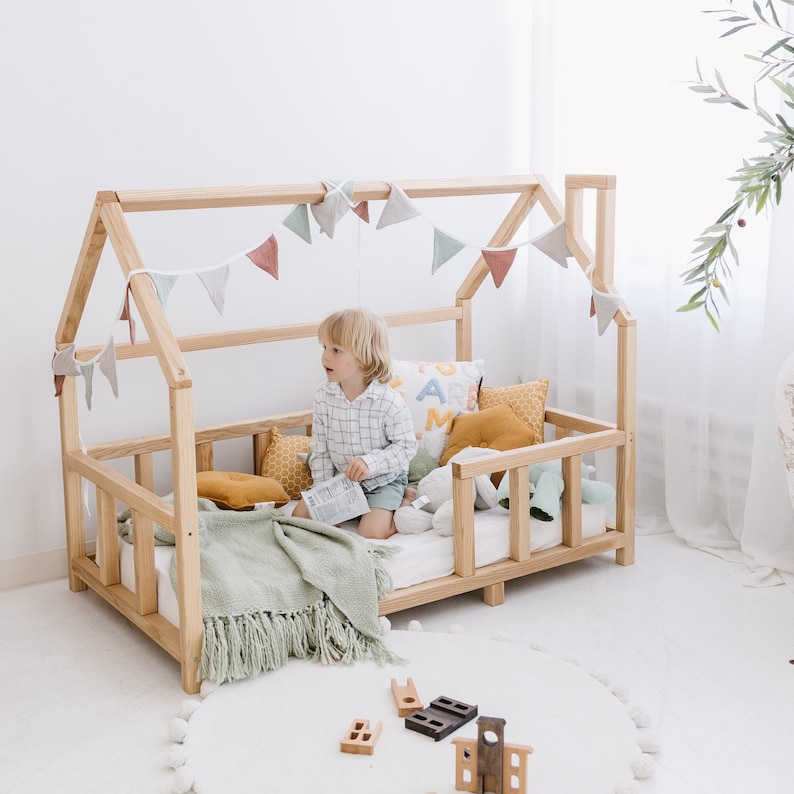 May include: A natural wood-framed toddler bed with a house-shaped frame. The bed is decorated with a garland of pastel-colored flags, pillows, and a green blanket. A young child sits inside the bed, surrounded by toys on a white rug.