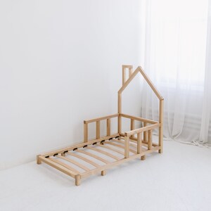 May include: A natural wood-toned toddler bed frame in a house shape. The bed frame has a slatted base and a low profile, with a house-shaped structure at the head of the bed. The bed is made of light-colored wood and is set against a white background.