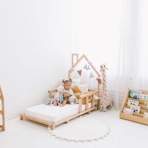 K&ouml;nnte beinhalten: Ein Kinderbett aus Holz mit einem hausf&ouml;rmigen Kopfteil und einer wei&szlig;en Matratze. Ein Kind sitzt auf dem Bett und liest ein Buch. Auf dem Bett befinden sich Stofftiere und Kissen. Auf der linken Seite des Bildes befindet sich ein B&uuml;cherregal aus Holz mit B&uuml;chern und auf der rechten Seite des Bildes ein B&uuml;cherregal aus Holz mit B&uuml;chern.