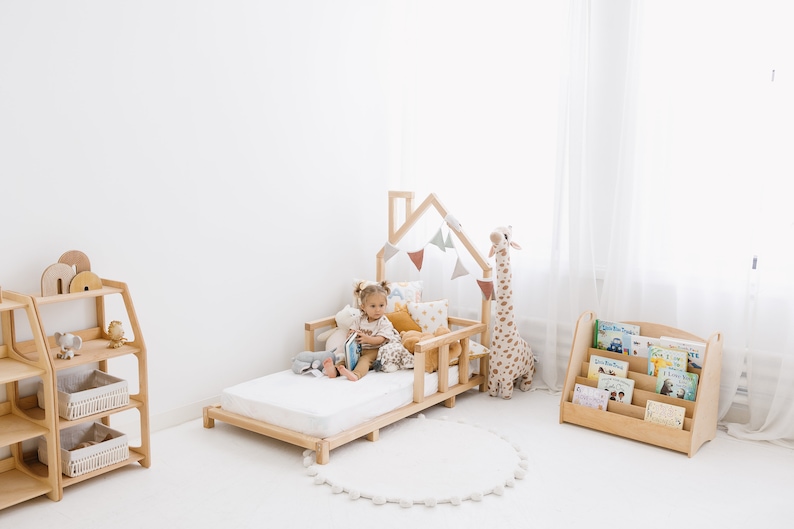 K&ouml;nnte beinhalten: Ein Kinderbett aus Holz mit einem hausf&ouml;rmigen Kopfteil und einer wei&szlig;en Matratze. Ein Kind sitzt auf dem Bett und liest ein Buch. Auf dem Bett befinden sich Stofftiere und Kissen. Auf der linken Seite des Bildes befindet sich ein B&uuml;cherregal aus Holz mit B&uuml;chern und auf der rechten Seite des Bildes ein B&uuml;cherregal aus Holz mit B&uuml;chern.