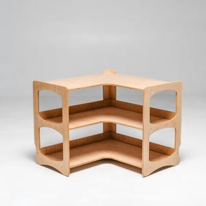 May include: A corner shelf unit made of light wood with three shelves. The shelves are open on all sides and have rounded edges. The unit is freestanding and has a simple, modern design.