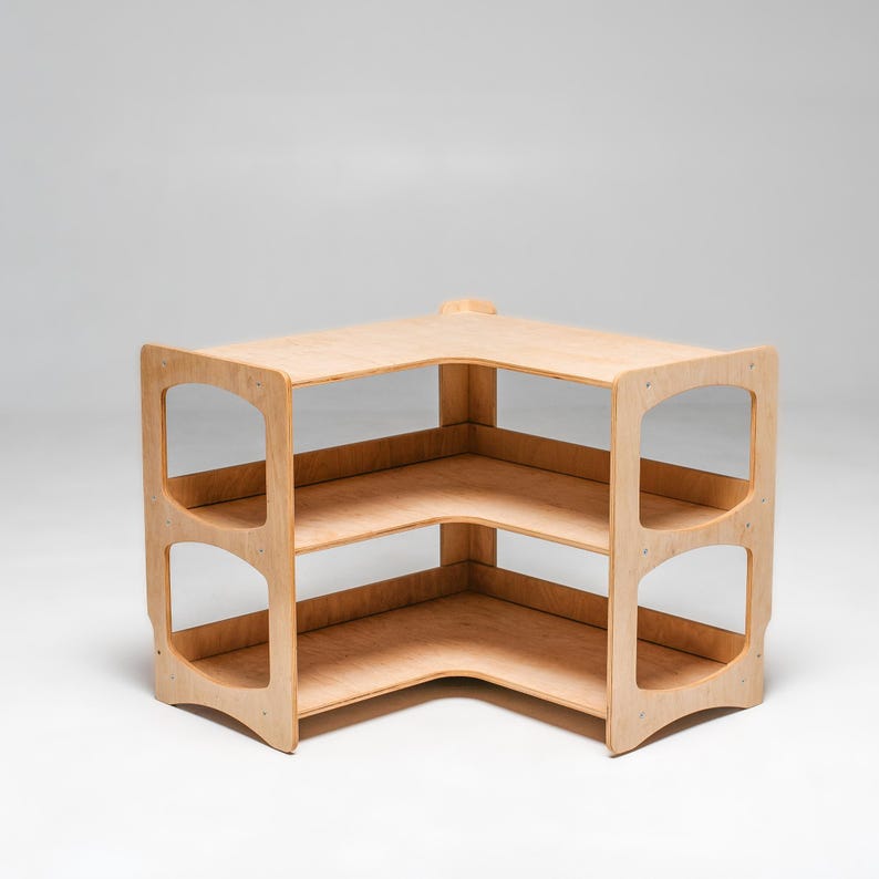 May include: A corner shelf unit made of light wood with three shelves. The shelves are open on all sides and have rounded edges. The unit is freestanding and has a simple, modern design.