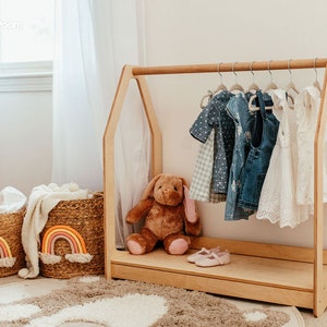May include: A light wood children's clothing rack with a house-shaped frame, holding various dresses and tops. Two woven baskets with rainbow accents sit nearby, along with a stuffed bunny and shoes. The room has a neutral color palette.