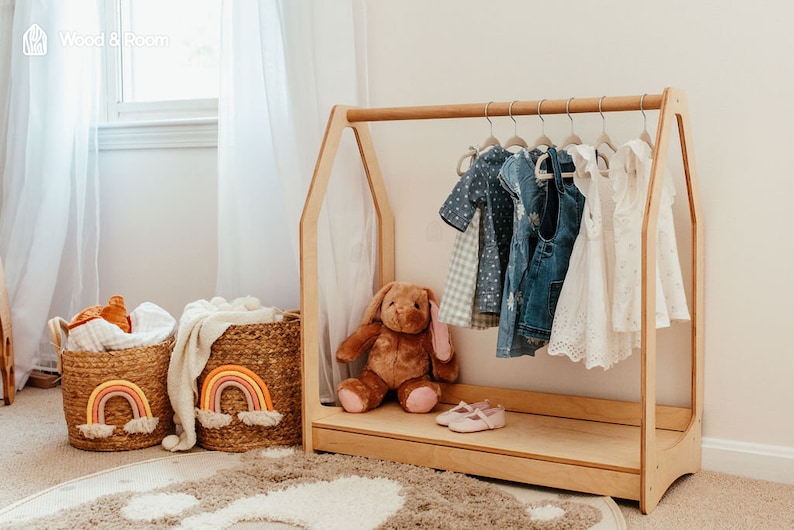 May include: A light wood children's clothing rack with a house-shaped frame, holding various dresses and tops. Two woven baskets with rainbow accents sit nearby, along with a stuffed bunny and shoes. The room has a neutral color palette.
