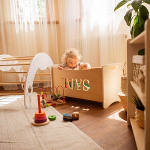 May include: A child's play area featuring a wooden toy box with the word "TOYS" cut out. Several wooden toys, such as a stacking ring and a bead maze, are on the floor. A white arch and climbing frame are in the background.
