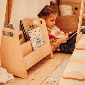 May include: A light wood bookshelf with four compartments holds a variety of children's books. A young girl sits on the floor reading a book.