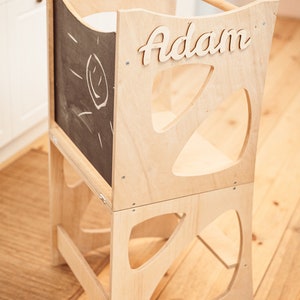 May include: A wooden learning tower with a chalkboard panel and the name "Adam" written on it. The tower has a cutout design and is made of natural wood.