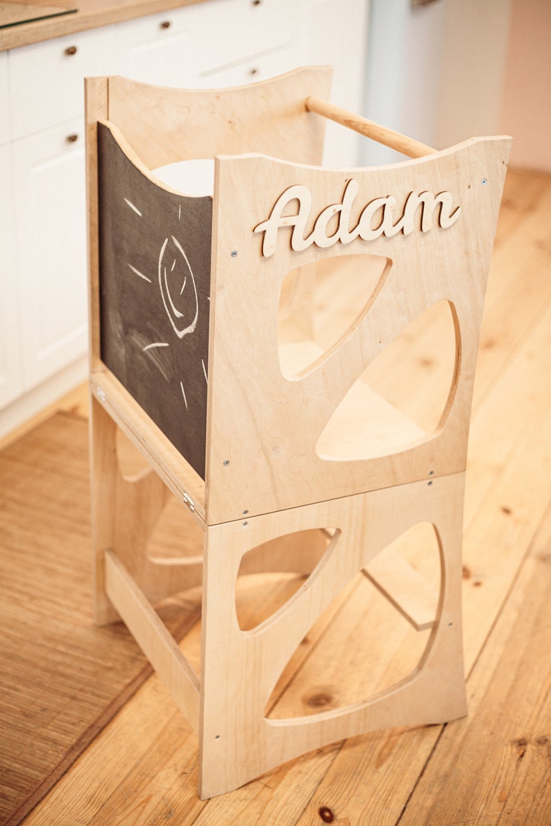 May include: A wooden learning tower with a chalkboard panel and the name "Adam" written on it. The tower has a cutout design and is made of natural wood.
