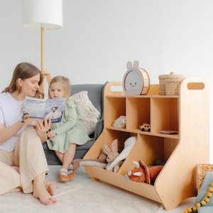 Puede incluir: Una unidad de almacenamiento de juguetes de madera con estantes y una cesta para juguetes. La unidad es de color marrón claro y tiene una luz nocturna con forma de conejo en el estante superior. Un niño está sentado en un sofá gris leyendo un libro titulado "God I Know You're There" a otro niño.