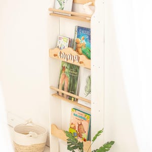 May include: White wooden bookshelf with three shelves, a wooden nameplate that says "emma", and a variety of children's books. The bookshelf is decorated with a small white rabbit figurine and a framed print with green leaves.
