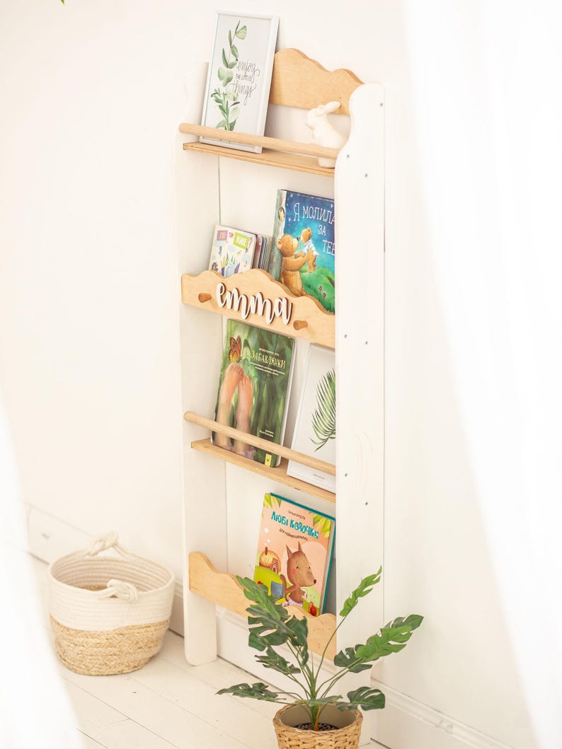 May include: White wooden bookshelf with three shelves, a wooden nameplate that says "emma", and a variety of children's books. The bookshelf is decorated with a small white rabbit figurine and a framed print with green leaves.