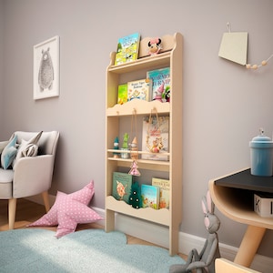 May include: A light-colored wooden bookshelf with four shelves, holding children's books and decorative items. The bookshelf has a scalloped top and sides, and is placed against a light gray wall. A pink star-shaped pillow rests on a light blue rug.