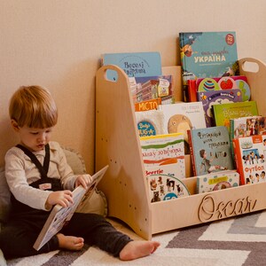 May include: A light wood bookshelf with a child's name "Oscar" engraved on the front. The bookshelf is filled with colorful children's books. A young child is sitting on the floor reading a book.