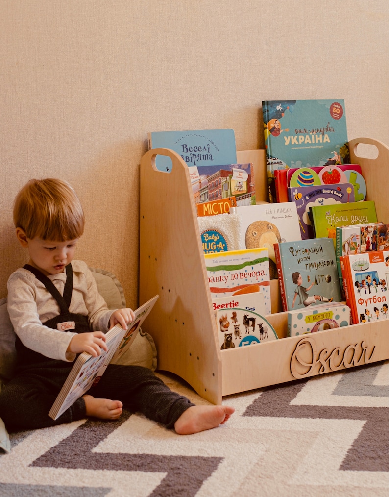 May include: A light wood bookshelf with a child's name "Oscar" engraved on the front. The bookshelf is filled with colorful children's books. A young child is sitting on the floor reading a book.
