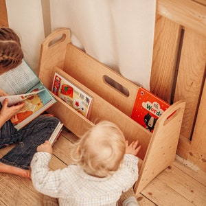 May include: Two children are sitting on a wooden floor, one reading a book and the other reaching into a wooden bookshelf with two shelves. The bookshelf has a handle on the top and a cutout handle on the side. There are several colorful books in the bookshelf.