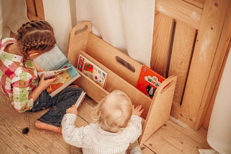 May include: Two children are sitting on a wooden floor, one reading a book and the other reaching into a wooden bookshelf with two shelves. The bookshelf has a handle on the top and a cutout handle on the side. There are several colorful books in the bookshelf.