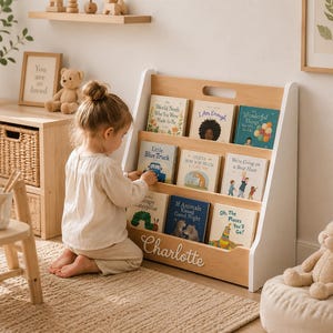 May include: A light wood and white floor-standing bookshelf featuring three slanted shelves filled with children's books. The front panel of the bookshelf is personalized with the name Charlotte in white script lettering and includes a cutout handle.