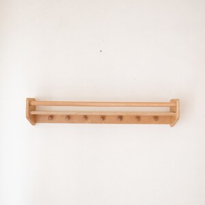 May include: A wooden wall-mounted shelf with a top rail and multiple pegs for hanging items. The shelf is made of light brown wood and has a natural finish.