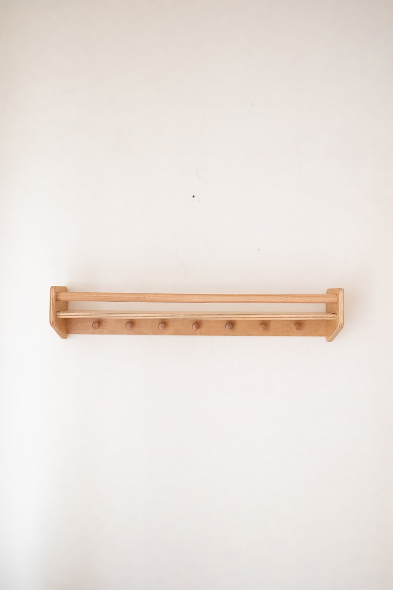 May include: A wooden wall-mounted shelf with a top rail and multiple pegs for hanging items. The shelf is made of light brown wood and has a natural finish.