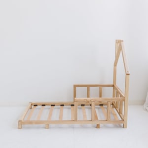 K&ouml;nnte beinhalten: Ein Kinderbettgestell aus Holz mit einem hausf&ouml;rmigen Kopfteil und einem Lattenrost. Das Bettgestell ist unbehandelt und hat eine nat&uuml;rliche Holzfarbe.
