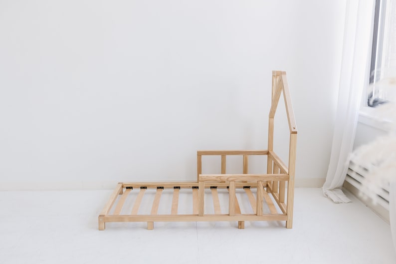 K&ouml;nnte beinhalten: Ein Kinderbettgestell aus Holz mit einem hausf&ouml;rmigen Kopfteil und einem Lattenrost. Das Bettgestell ist unbehandelt und hat eine nat&uuml;rliche Holzfarbe.