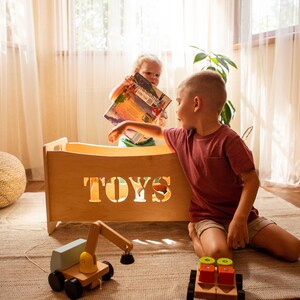 May include: A wooden toy chest with the word "TOYS" cut out, and two children playing with toys. A wooden toy excavator and a wooden toy truck are on the floor. The room has a neutral colour palette.