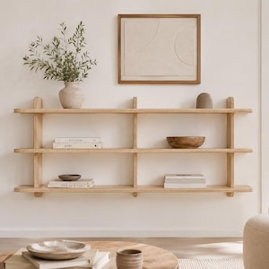 May include: A three-tiered light wood wall shelf with vertical supports. The shelves hold a ceramic vase with greenery, a wooden bowl, and several stacked books. Above the shelf hangs a framed piece of art with a neutral geometric design.