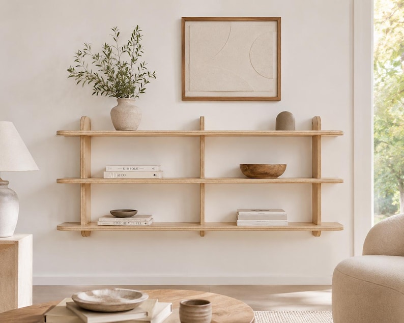 May include: A three-tiered light wood wall shelf with vertical supports. The shelves hold a ceramic vase with greenery, a wooden bowl, and several stacked books. Above the shelf hangs a framed piece of art with a neutral geometric design.