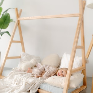 May include: A wooden teepee bed frame with a white blanket, pillows, and a child lying on it. The child is wearing a plaid pajama top and holding a stuffed animal.