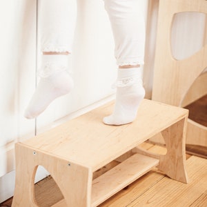 May include: A light wood step stool with a curved design and a shelf. The stool is made of natural wood and has a light brown finish. The stool is designed for children to reach higher surfaces.