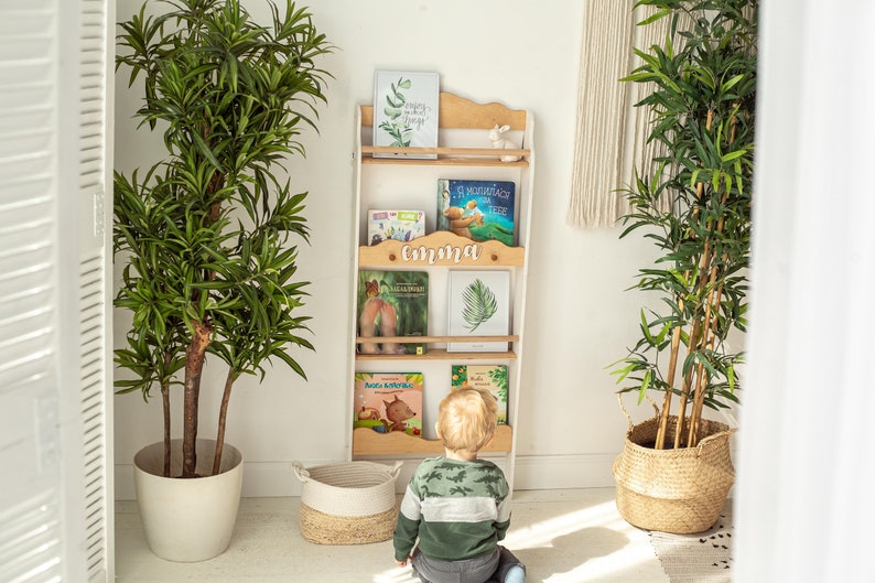 May include: A white and wood-toned bookshelf with books and the name "Emma" on it. The bookshelf is leaning against a white wall, with two large potted plants on either side. A child is looking at the bookshelf.