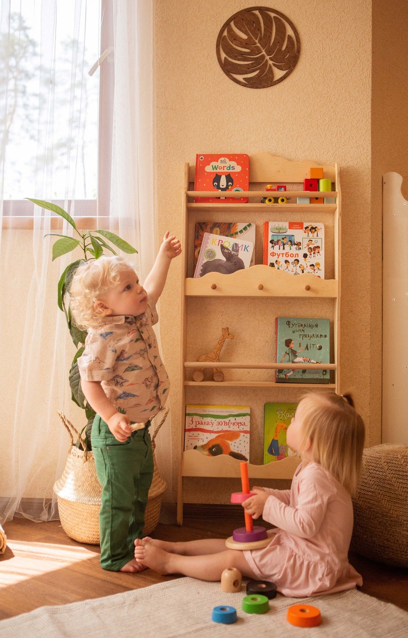 May include: A light wooden bookshelf filled with colorful children's books. Two young children are in the room, one pointing at the shelf. A wooden giraffe toy sits on a shelf. A decorative leaf hangs on the wall.