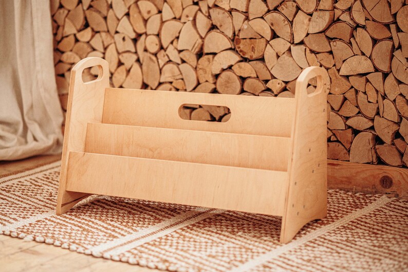 Puede incluir: Una estanter&iacute;a de madera clara con tres estantes y dos asas. La estanter&iacute;a est&aacute; sobre una alfombra de color marr&oacute;n y blanco con un patr&oacute;n.