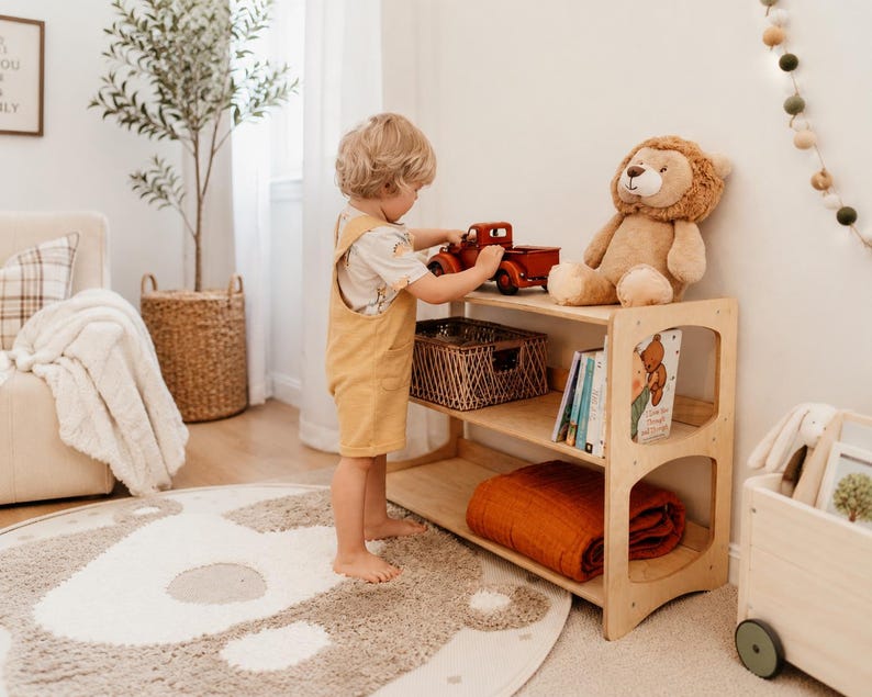 May include: A light-colored wooden bookshelf with three tiers, featuring a red toy truck and a plush lion on the top shelf, a woven basket on the middle shelf, and a folded rust-colored blanket on the bottom shelf alongside several books.