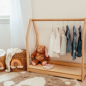 May include: A light-colored wooden A-frame clothing rack with a horizontal bar holding six small garments on hangers. A brown plush rabbit sits on the base next to a pair of light pink shoes. Two woven baskets with rainbow designs sit nearby.