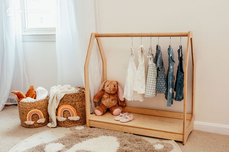 May include: A light-colored wooden A-frame clothing rack with a horizontal bar holding six small garments on hangers. A brown plush rabbit sits on the base next to a pair of light pink shoes. Two woven baskets with rainbow designs sit nearby.