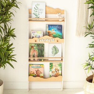 May include: White wooden bookshelf with five shelves, featuring a variety of children's books. The top shelf has a framed print with the text "Enjoy the little things". The second shelf has a book with the text "Я молилась за тебя". The third shelf has a book with the text "Забавники!" and a book with the text "Emma". The fourth shelf has a book with the text "Люби котиков" and a book with the text "Хватит бояться".