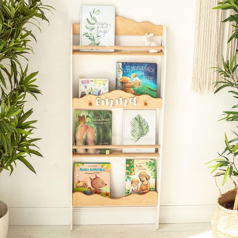 May include: White wooden bookshelf with five shelves, featuring a variety of children's books. The top shelf has a framed print with the text "Enjoy the little things". The second shelf has a book with the text "Я молилась за тебя". The third shelf has a book with the text "Забавники!" and a book with the text "Emma". The fourth shelf has a book with the text "Люби котиков" and a book with the text "Хватит бояться".