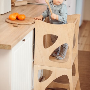 May include: A light wood toddler learning tower with a child standing on the top platform holding a wooden spoon. The tower has two platforms and a cutout design. The child is wearing a blue shirt and grey pants.