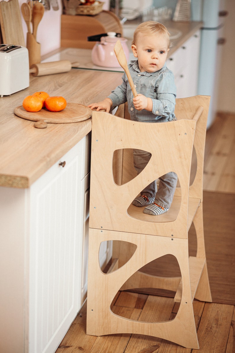 May include: A light wood toddler learning tower with a child standing on the top platform holding a wooden spoon. The tower has two platforms and a cutout design. The child is wearing a blue shirt and grey pants.