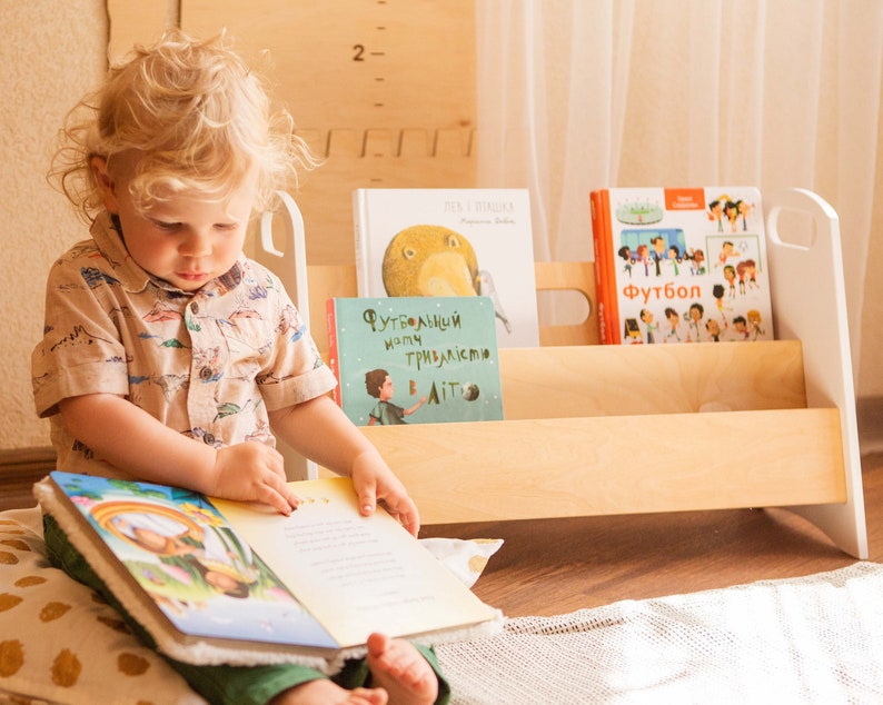 May include: A young child sits on a floor with a colorful book open in their lap. The child is wearing a light brown and white patterned shirt and green pants. The child is looking at the book. Behind the child is a wooden bookshelf with three shelves. The shelves are filled with books. The books have colorful covers with Russian text. The bookshelf is made of light wood and has a white painted finish.