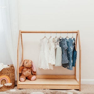 May include: A light-colored wooden A-frame clothing rack featuring a horizontal hanging rod. Several children's garments are suspended on hangers, including white dresses, a checkered top, and blue patterned outfits. A brown plush rabbit sits inside.
