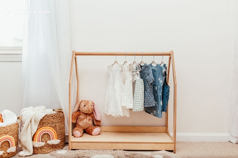 May include: A light-colored wooden A-frame clothing rack featuring a horizontal hanging rod. Several children's garments are suspended on hangers, including white dresses, a checkered top, and blue patterned outfits. A brown plush rabbit sits inside.