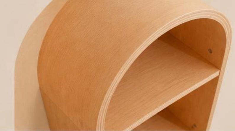 May include: A light brown, curved wooden shelf unit with two shelves. The unit has a rounded top and sides, and the wood grain is visible. The shelves are open and provide storage space. The design is modern and minimalist.