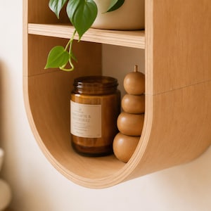 May include: A light brown, curved wooden shelf with two levels. The top shelf holds a potted plant. The lower shelf displays a brown glass jar candle and a stack of wooden spheres. The candle label reads "Bergamot & Neroli."