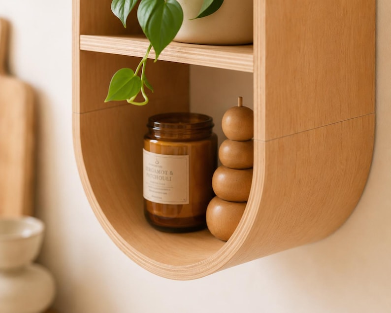 May include: A light brown, curved wooden shelf with two levels. The top shelf holds a potted plant. The lower shelf displays a brown glass jar candle and a stack of wooden spheres. The candle label reads "Bergamot & Neroli."