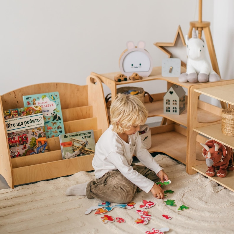 May include: A young child sits on a white rug playing with a colorful puzzle. The puzzle pieces are shaped like animals and are scattered around the child. The child is wearing a white shirt and brown pants. The child is sitting in front of a wooden bookshelf with books on the shelves. The bookshelf is next to a wooden shelf with a white unicorn stuffed animal on top.