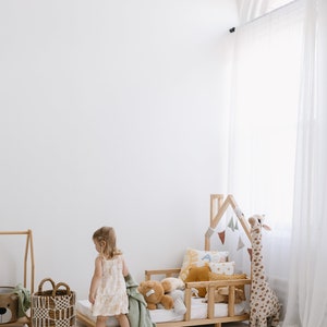 K&ouml;nnte beinhalten: Ein kleines Kind steht in der N&auml;he eines Kinderbettes aus Holz mit einer wei&szlig;en und gr&uuml;nen Decke, einer Stoffgiraffe und verschiedenen bunten Kissen. Das Bett ist mit einem h&ouml;lzernen, hausf&ouml;rmigen Kopfteil und einem wei&szlig;en Pompon-Teppich verziert.